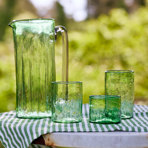 A set of six Viento Small Green Tumblers by Caskata, crafted from recycled glass for unique character, are arranged outdoors on a striped cloth with blurred greenery in the background.