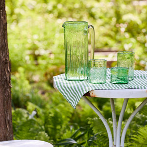 A set of six Viento Small Green Tumblers by Caskata is displayed on a round white table with a green-striped cloth in a lush garden setting.