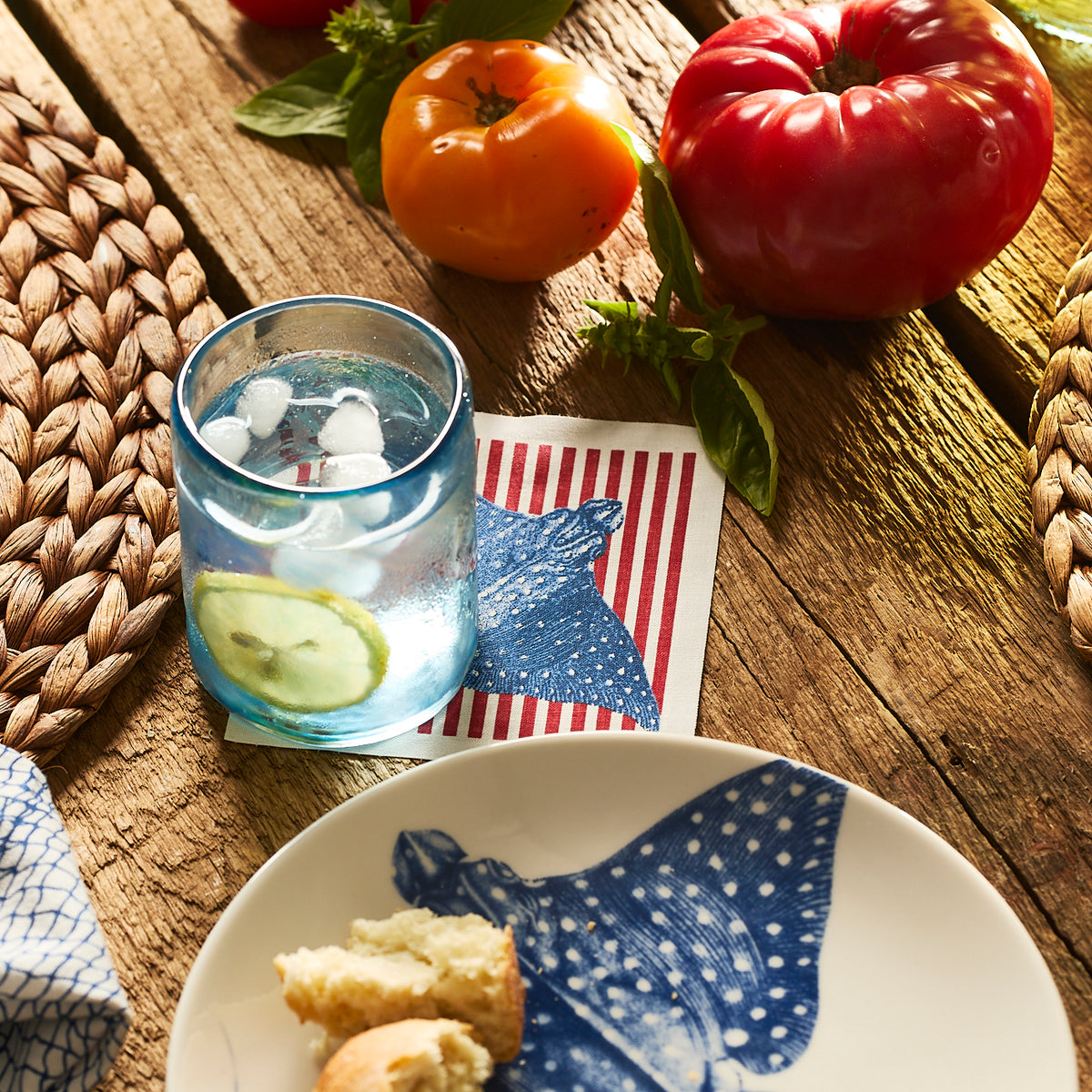 A glass of iced water with lemon on a coaster sits beside tomatoes, basil, and a partially eaten roll on a wooden table featuring the MY DRAP Seaside Stripes Cocktail Napkin Roll.