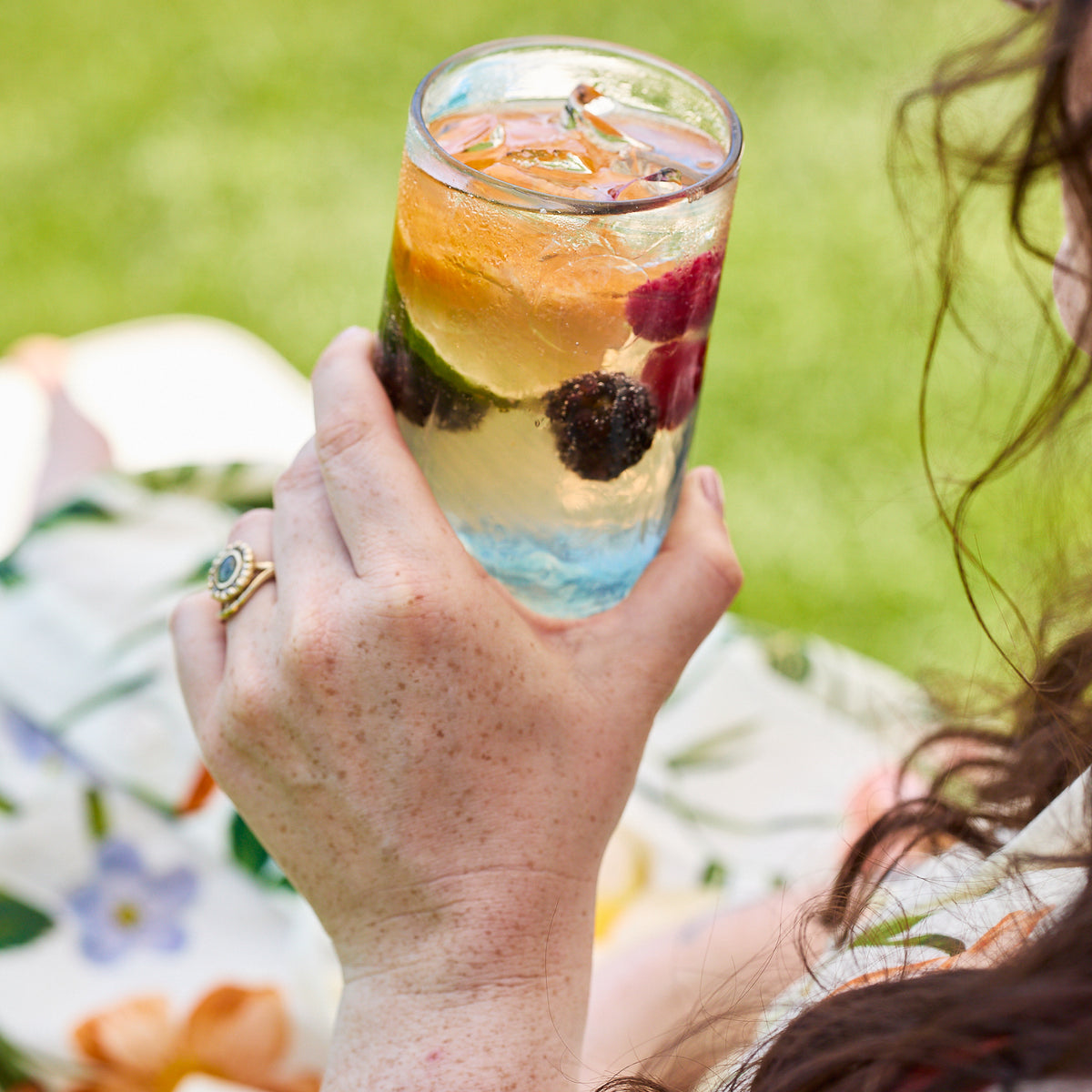 A person holds a glass of sparkling water with ice, orange slice, blackberries, and raspberries in a Caskata Viento Large Blue Tumbler (set of 6); floral dress and green grass in the background.