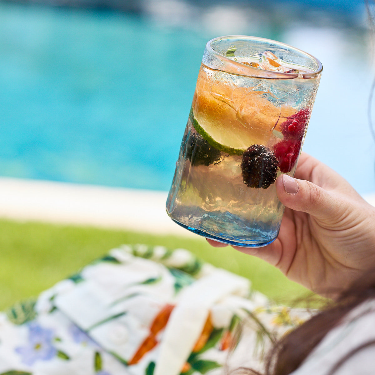 A person holds a Caskata Viento Large Blue Tumbler with a mixed drink by the pool, floral shirt visible. This handblown set of 6 highlights beautiful recycled glass craftsmanship.