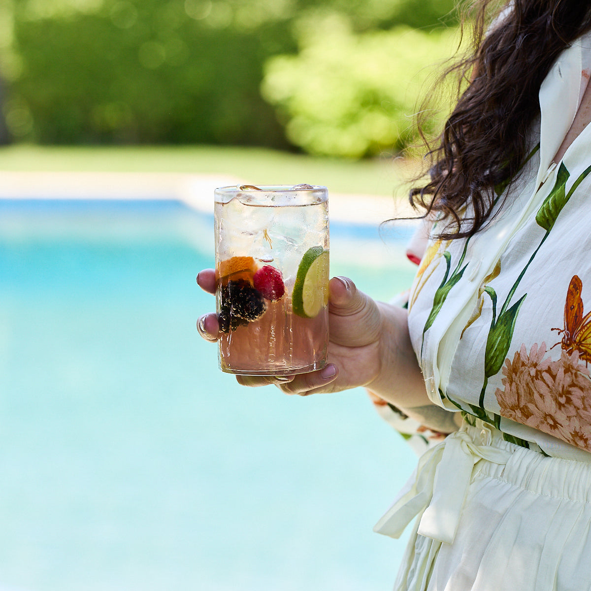 By the pool, someone in floral attire holds a glass of iced fruit drink with berries and lime, served in a Caskata Viento Large Clear Tumbler from the set of 6.