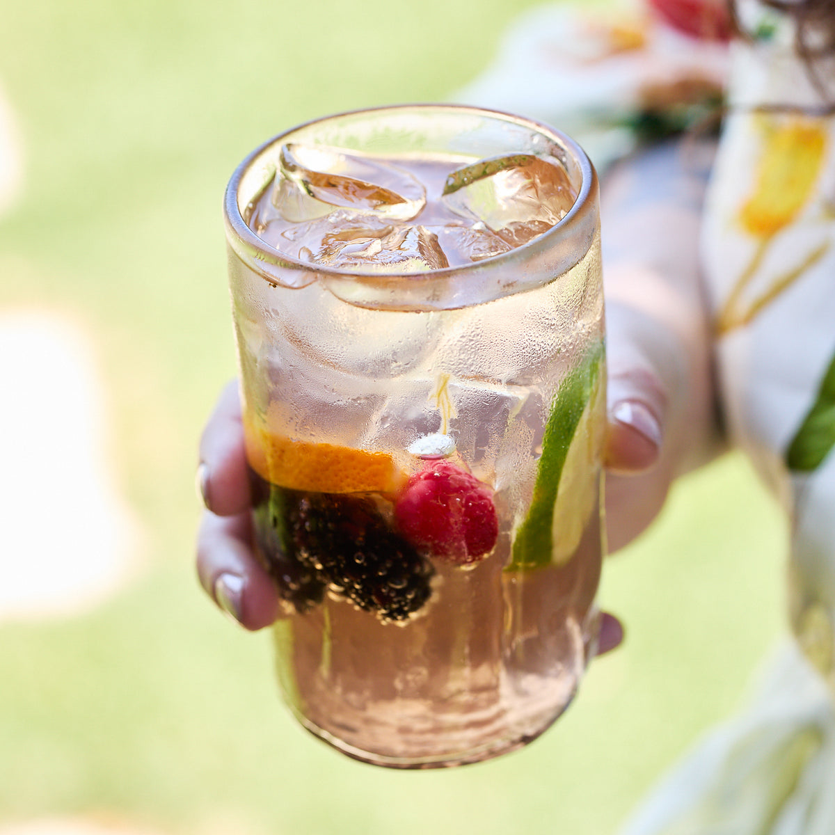 A hand holds a glass of iced clear beverage with lime, orange slice, cherry, and blackberry in beautiful recycled glassware from the Caskata "Viento Essentials" Serving Bundle.