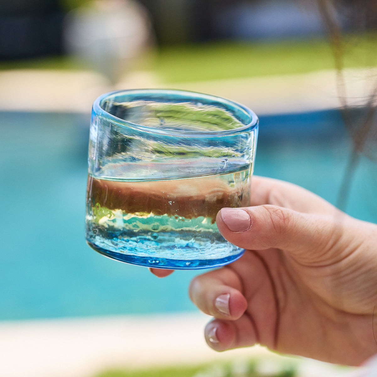 A hand holds a Caskata Viento Small Blue Tumbler filled with clear liquid near a shimmering pool, the background softly blurred. Set of 6.
