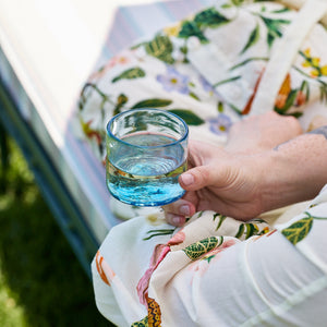 A person holds a Caskata Viento Small Blue Tumbler from the set of 6 while sitting outdoors, showcasing its handblown recycled glass.