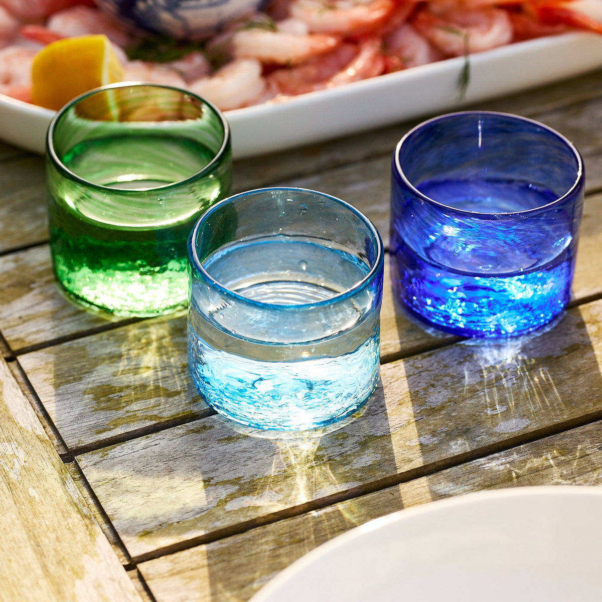 Three Viento Small Green Tumblers by Caskata, handblown from recycled glass and filled with water, are set on a wooden table beside a white platter of seafood.