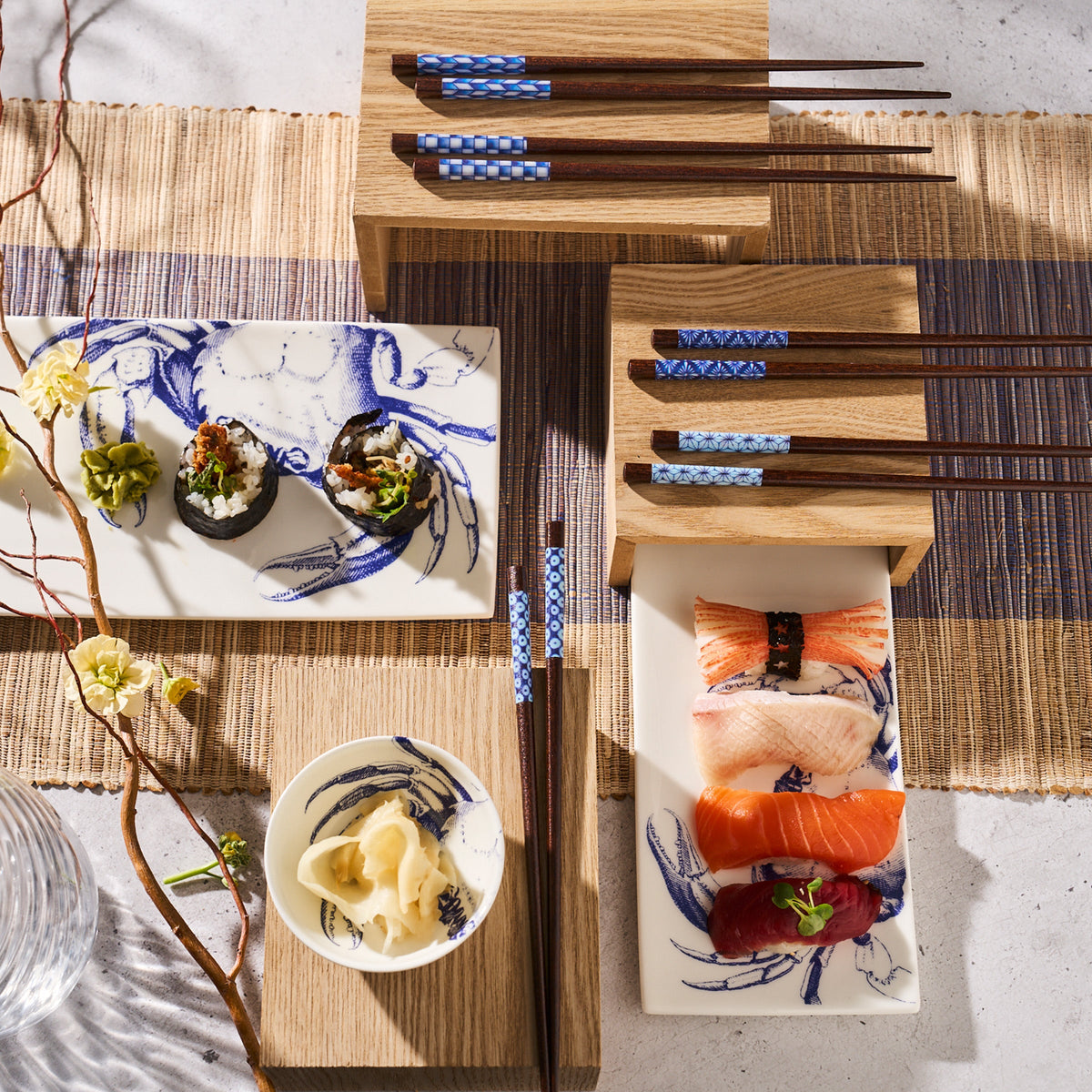 A Japanese meal setup with sushi, sashimi, pickled ginger, and hand rolls on high-style ceramic plates and Caskata Crab Medium Sushi Trays, Set of 2. Wooden stands and a mat complete the elegant arrangement. Chopsticks with blue designs featuring crab patterns are placed beside all dishes.