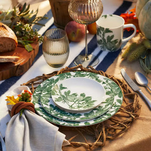 The Caskata Arbor Green Table for 4 features leaf-patterned dinnerware, a mug, glass, bread, apple, and a yellow flower napkin on a rustic outdoor table.
