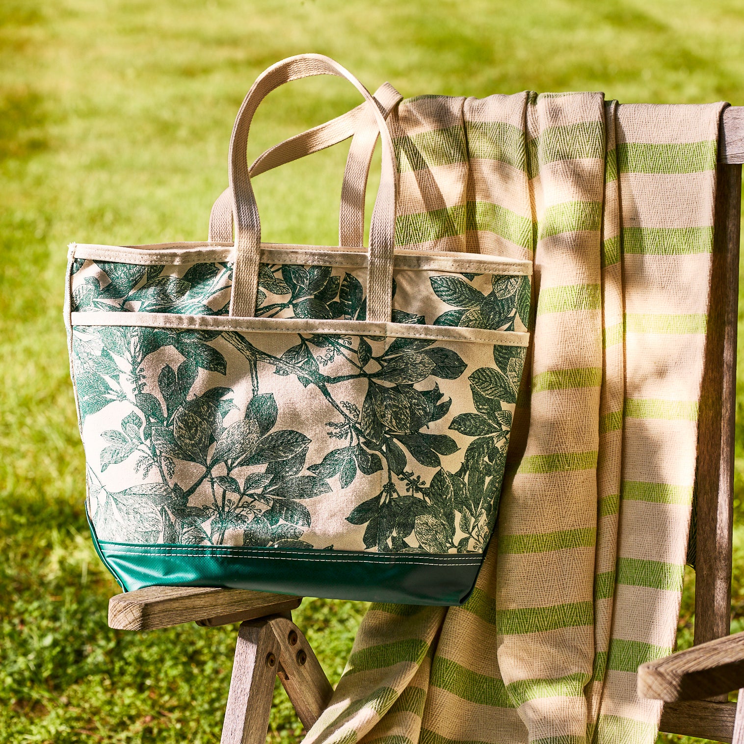 The Caskata x Steele Everyday Tote Bag Arbor Green, handcrafted in the USA, features a green botanical print and sits atop a striped beige and green blanket on a wooden chair outdoors.