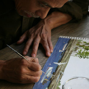 A person is meticulously painting a detailed waterfront scene on a sketchpad, concentrating intensely on their work, much like the serene artistry found in Caskata's By the Sea Mugs, Set of 4 by Felix Doolittle.