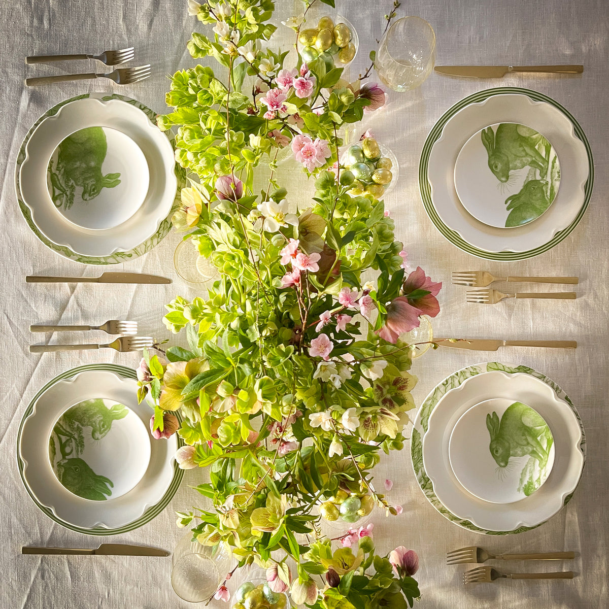 A table setting with four Bunnies Verde Small Plates by Caskata featuring botanical designs, placed around a lush green floral centerpiece. Cutlery and glasses are neatly arranged around each plate on a white tablecloth.