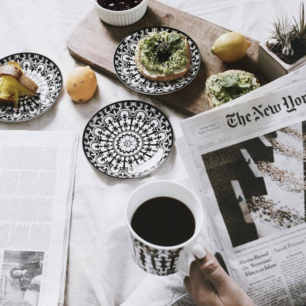 Person holding a coffee cup and reading a newspaper, with avocado toast on patterned Casablanca Small Plates from Caskata Artisanal Home, lemons, and a bowl of cherries on the table.