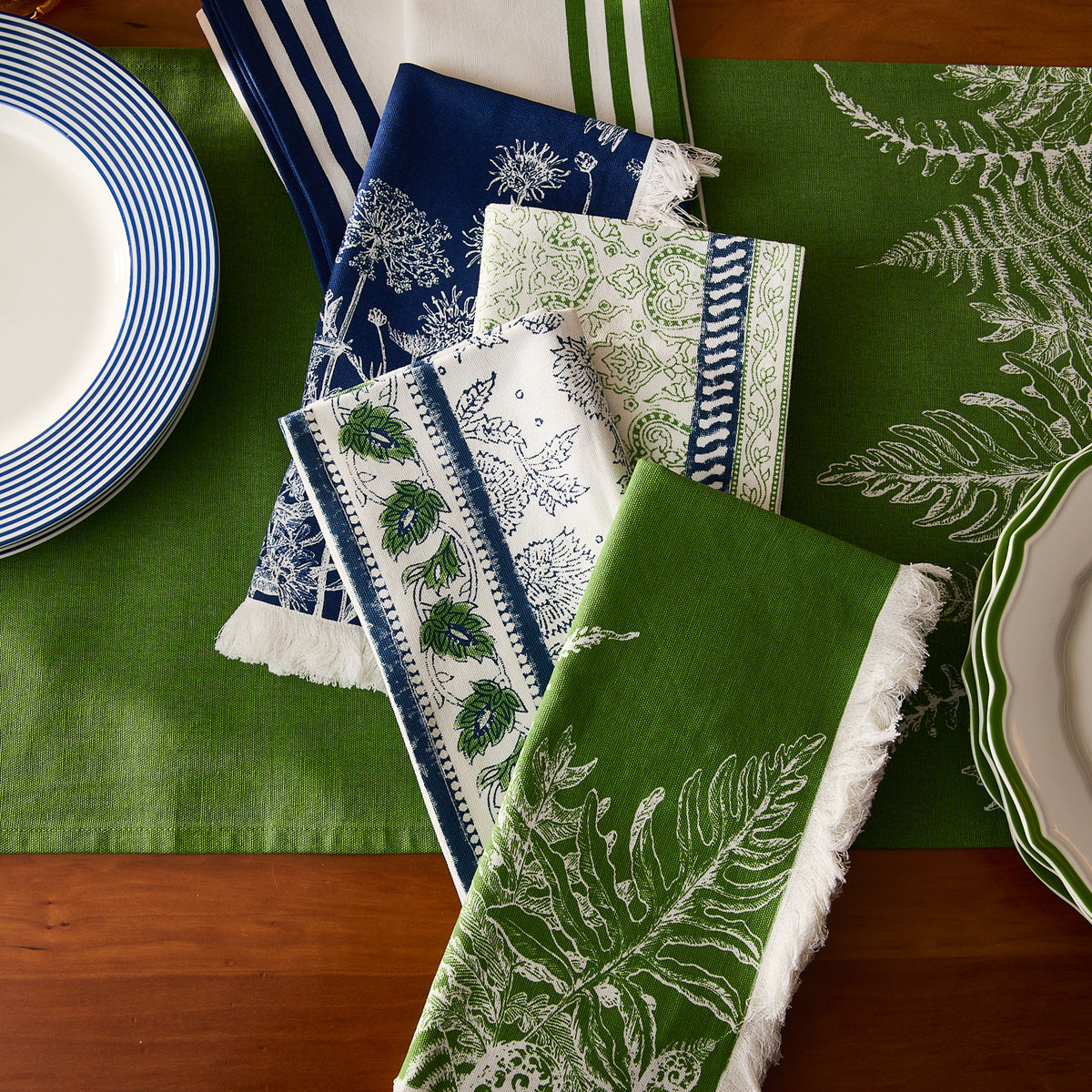 Caskata Block Island Block Print Dinner Napkins, in blue, white, and green, paired with plates on a green table runner add bohemian flair to a wooden table.