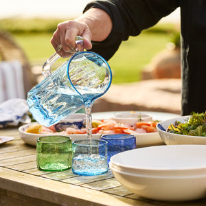 A person pours water from the Caskata Viento Large Blue Pitcher into a glass on an outdoor table set with food, bowls, and glassware.