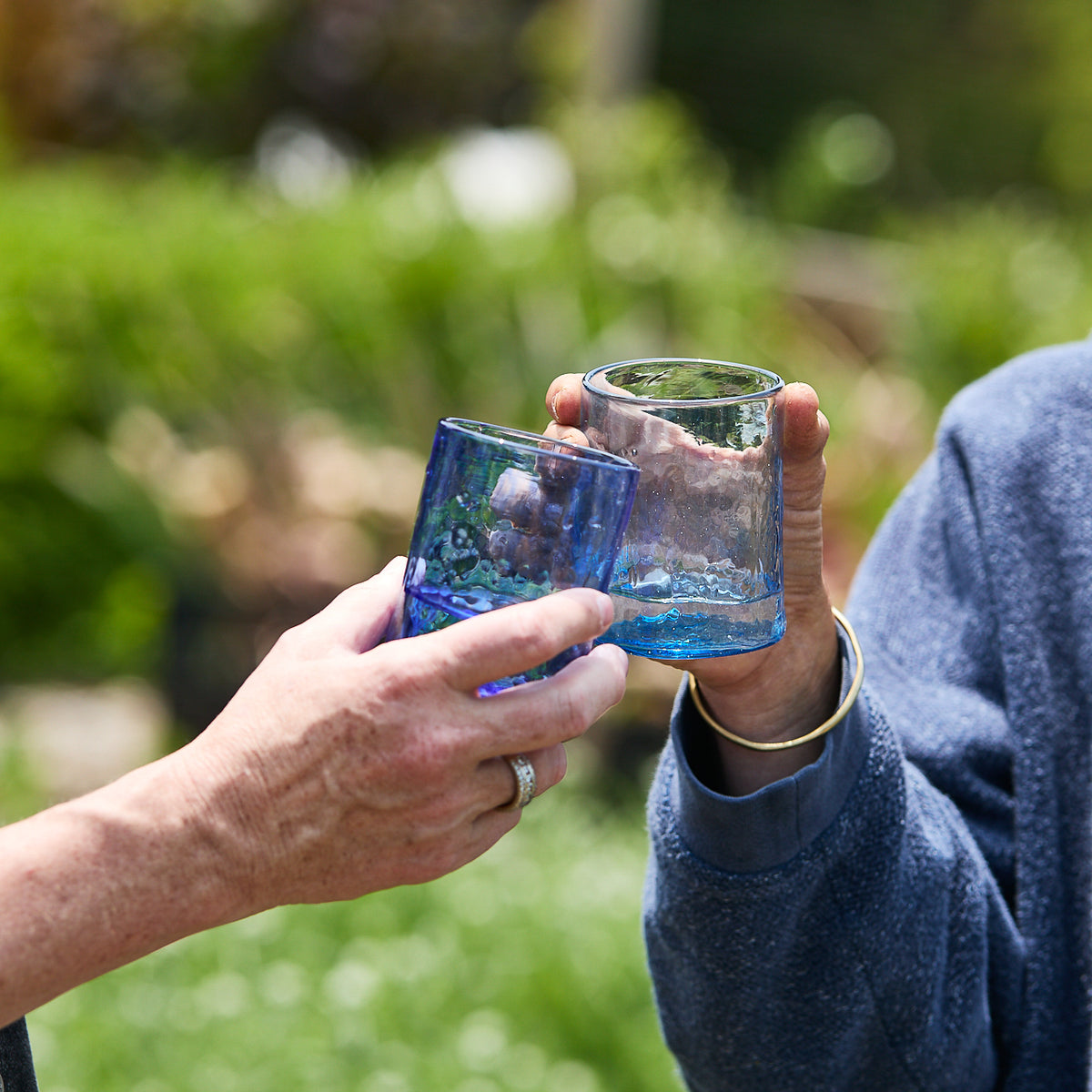 Two people clink Caskata Viento Medium Blue Tumblers from a set of 6 together outdoors, with greenery blurred in the background.