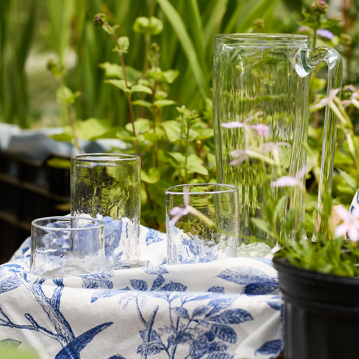 The Caskata "Viento Essentials" Serving Bundle, with handblown recycled glass tumblers and pitcher, rests on a white and blue floral cloth outdoors amid lush green plants and flowers.