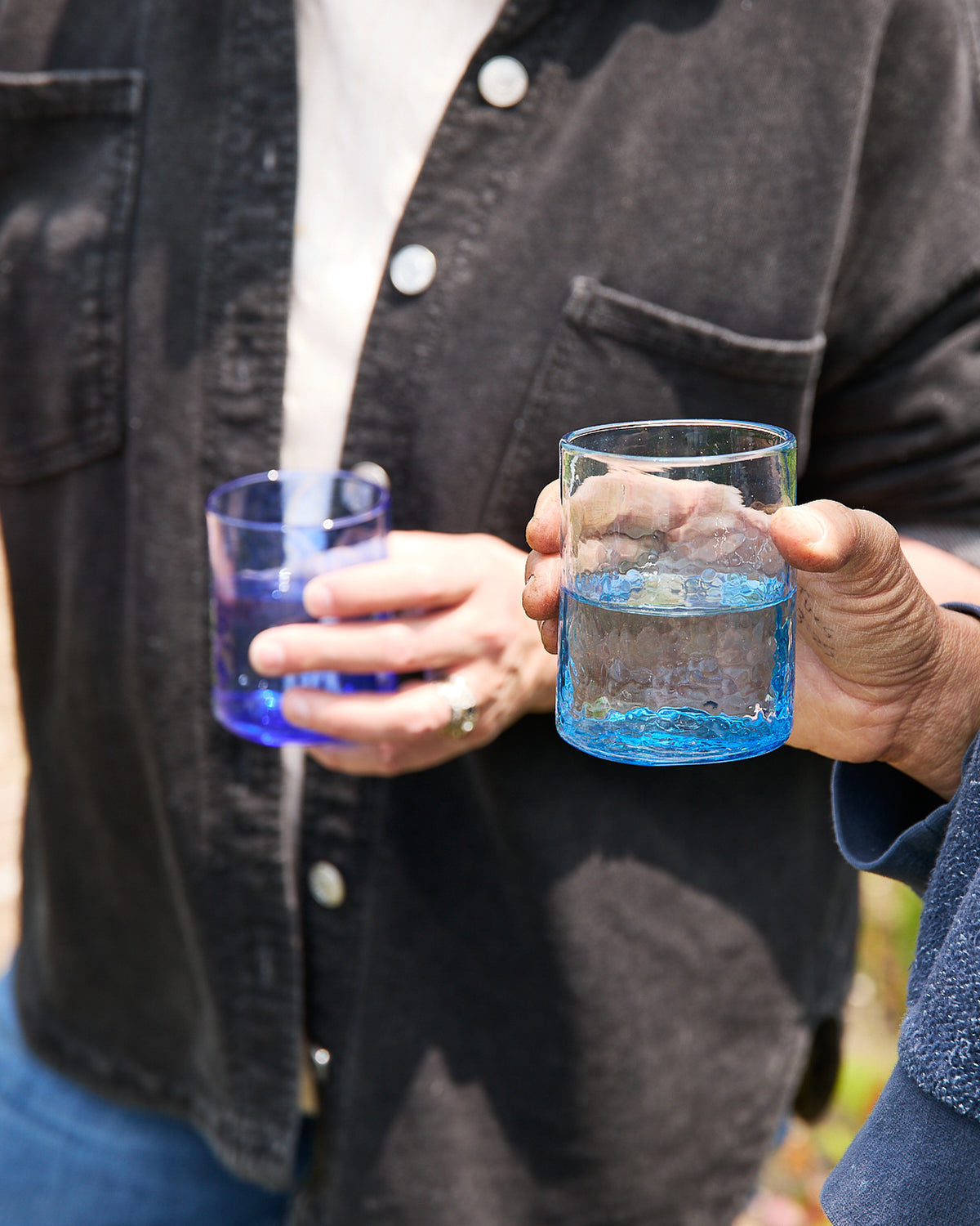 Two people hold Caskata’s Viento Medium Blue Tumblers—a set of 6 handblown pieces made from recycled glass—each featuring elegant designs and a unique blue tint.