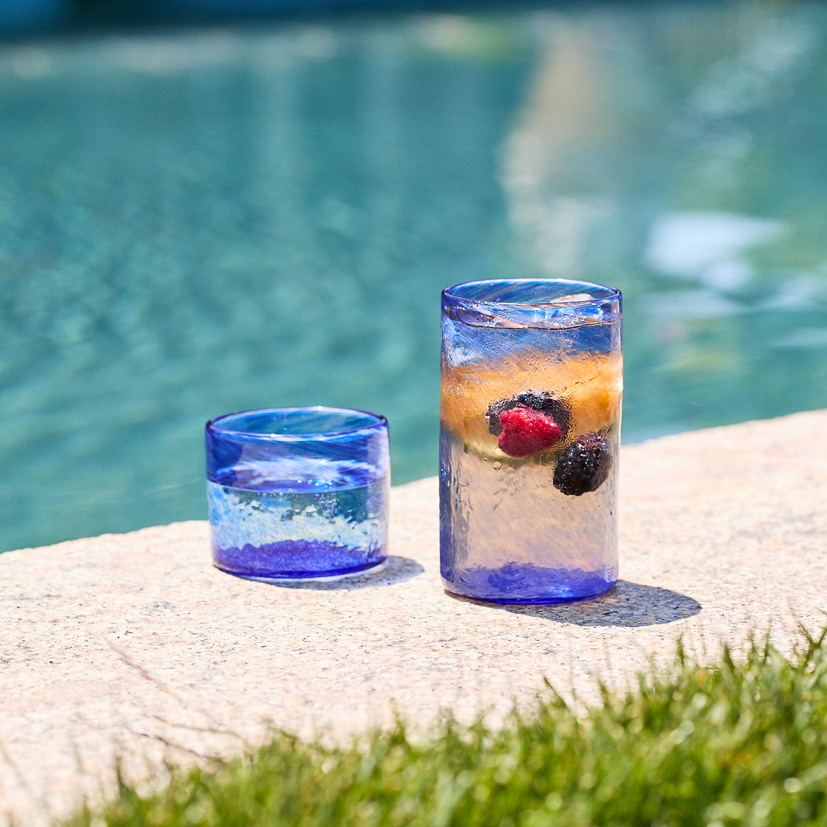 Two Caskata Viento Large Cobalt Tumblers from the set of 6 sit poolside; one is empty while the other holds water with floating berries and an orange slice.