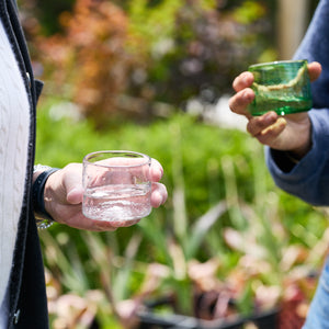 Two people stand outdoors holding small glass cups—one clear with water, the other a Caskata Viento Small Green Tumbler—both handmade from recycled glass, with lush plants visible in the background.