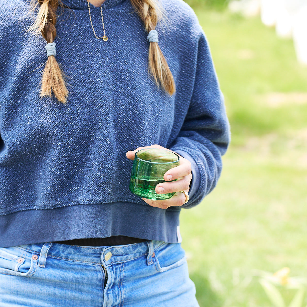 A person in a blue sweater and jeans holds a Caskata Viento Small Green Tumbler outdoors, with braided hair and a gold necklace visible.
