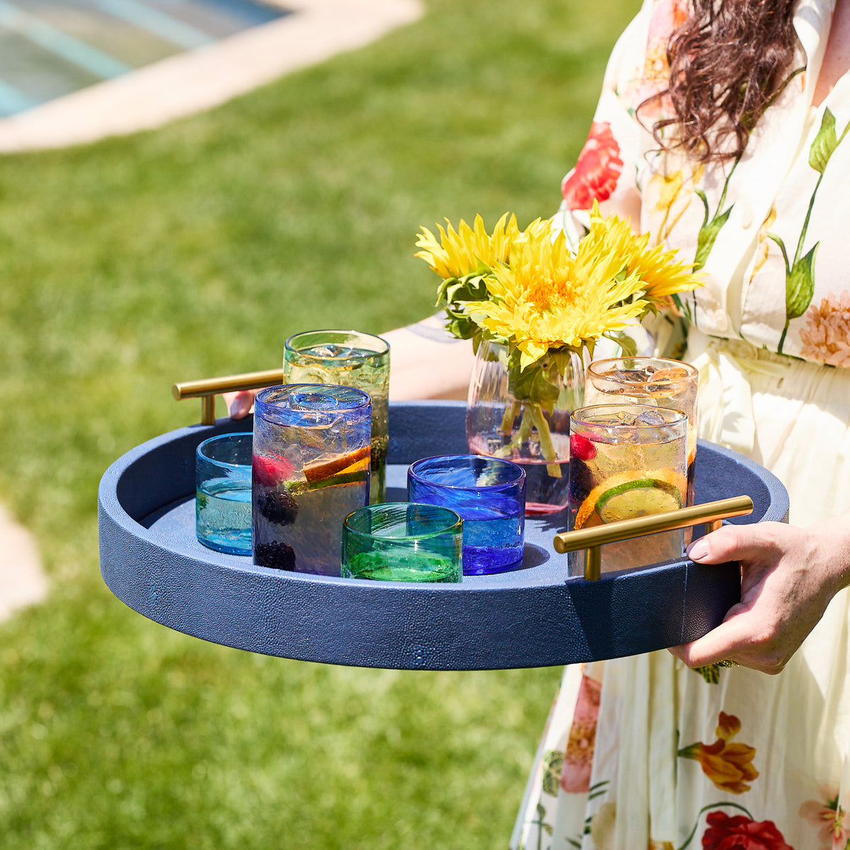 On a grassy lawn, someone in a floral dress carries a round blue tray with Caskata’s Viento Small Blue Tumblers, a glass vase of yellow flowers, and fruit-infused drinks.