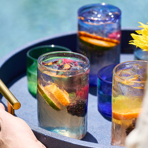 A tray holds glasses of sparkling water with citrus and berries alongside Caskata’s Viento Large Blue Tumblers, set of 6, crafted from recycled glass and gleaming by the pool.