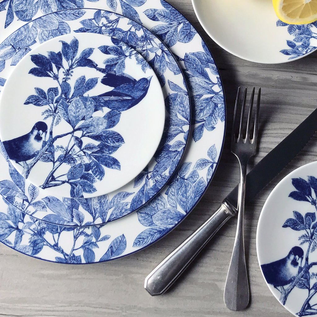 A table setting with heirloom-quality Arbor Blue Birds Small Plates by Caskata Artisanal Home featuring bird and leaf patterns, accompanied by a fork and knife, and a partially visible lemon slice on a small plate. The botanical details add an elegant touch to the scene.
