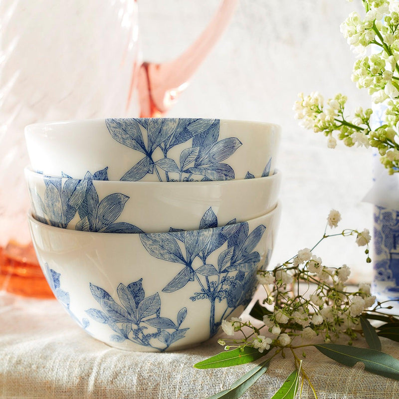 Three premium Caskata Arbor Cereal Bowls with blue botanical patterns are stacked together, alongside a bouquet of small white flowers and a clear pink pitcher in the background.