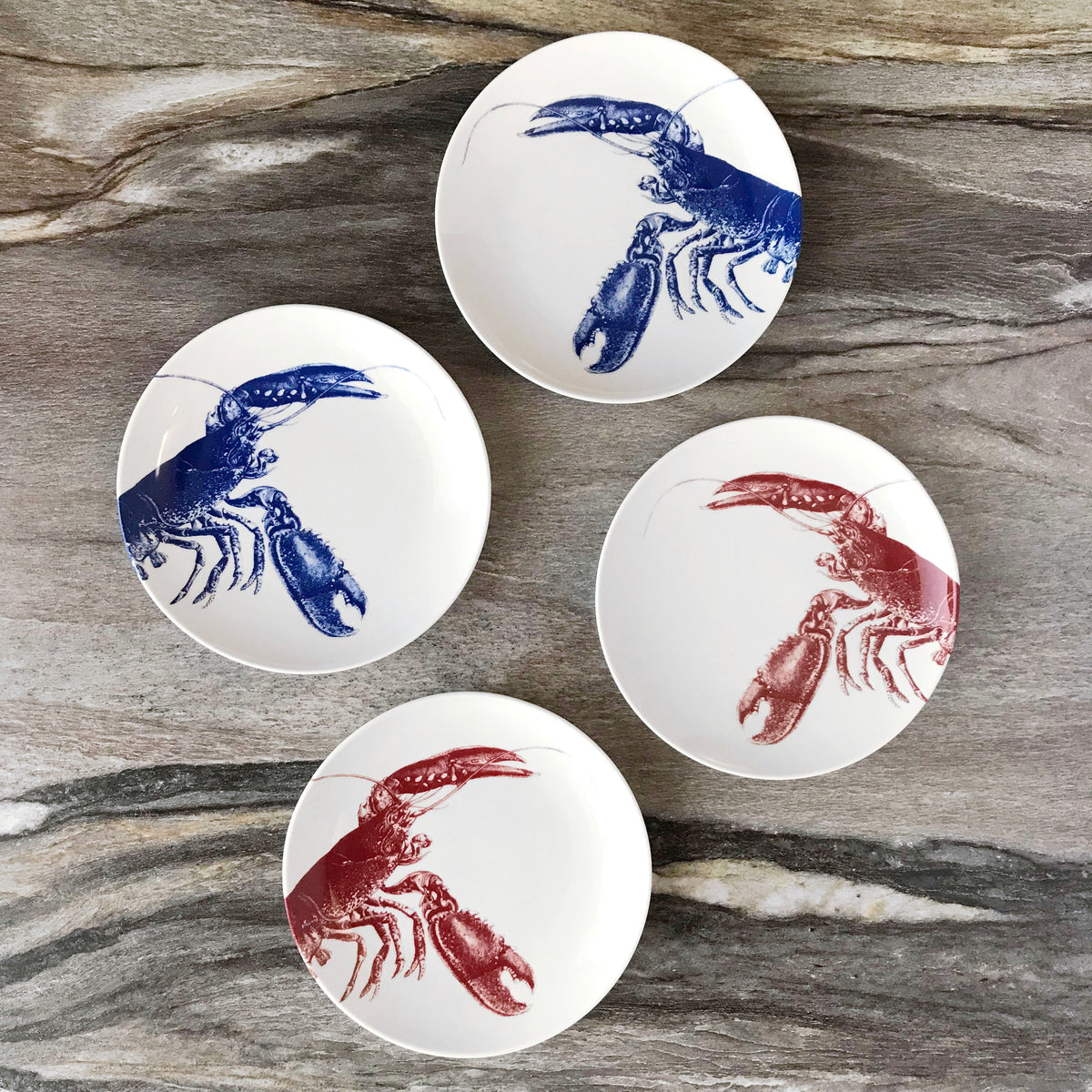 Four heirloom-quality Lobster Small Plates by Caskata Artisanal Home on a gray-marble background, each featuring a blue or red illustration of a lobster, reminiscent of New England&#39;s coast.