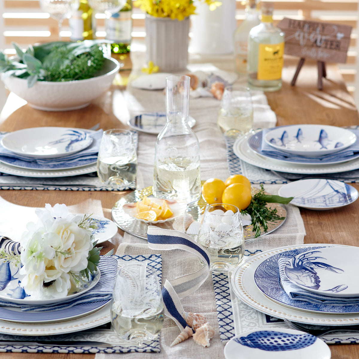 A dining table set with heirloom-quality dinnerware featuring a crab pattern, blue and white dishware, lemons, Caskata Artisanal Home Crab Small Plates, a carafe of water, decorative flowers, and various condiments. There is a centerpiece and a background with bottles and flowers.
