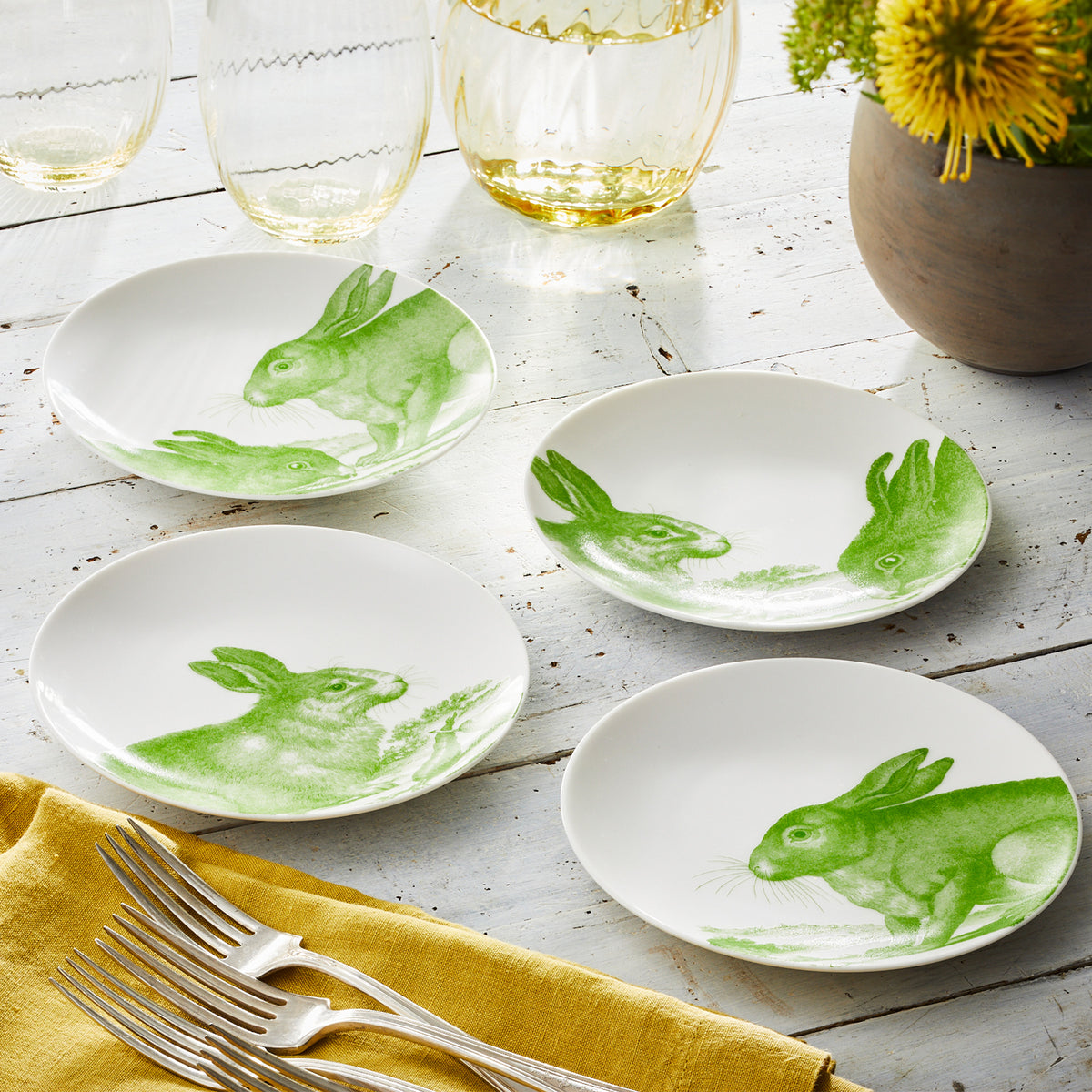 Four high-fired Caskata Bunnies Verde Small Plates with green bunny scene illustrations placed on a wooden table. Nearby are drinking glasses, a bunch of yellow flowers in a pot, a yellow napkin, and several forks.
