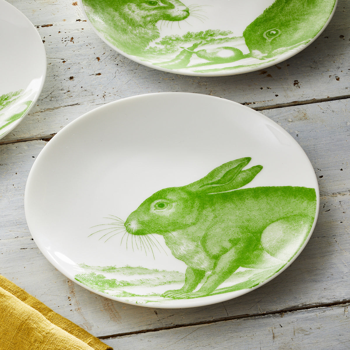 A Caskata Bunnies Verde Small Plate featuring a green illustration of a bunny scene, sitting on a light gray wooden surface with a yellow cloth nearby.