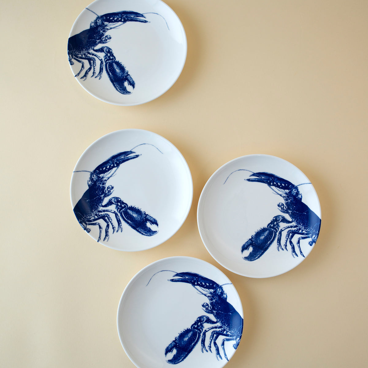 Four small plates with blue lobster designs are arranged in a diagonal line on a beige background, capturing the essence of New England&#39;s coast. These **Lobster Small Plates** from **Caskata Artisanal Home** bring a touch of seaside charm to any setting.