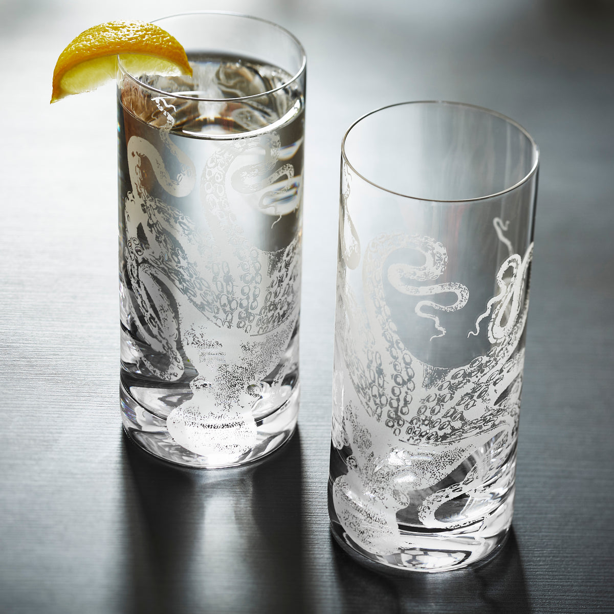 Lucy the Octopus Tall Cocktail Glasses sold as a set of 2 in hand-etched lead-free crystal barware from Caskata. One is shown full of water and garnished with a slice of lemon, the other is empty.