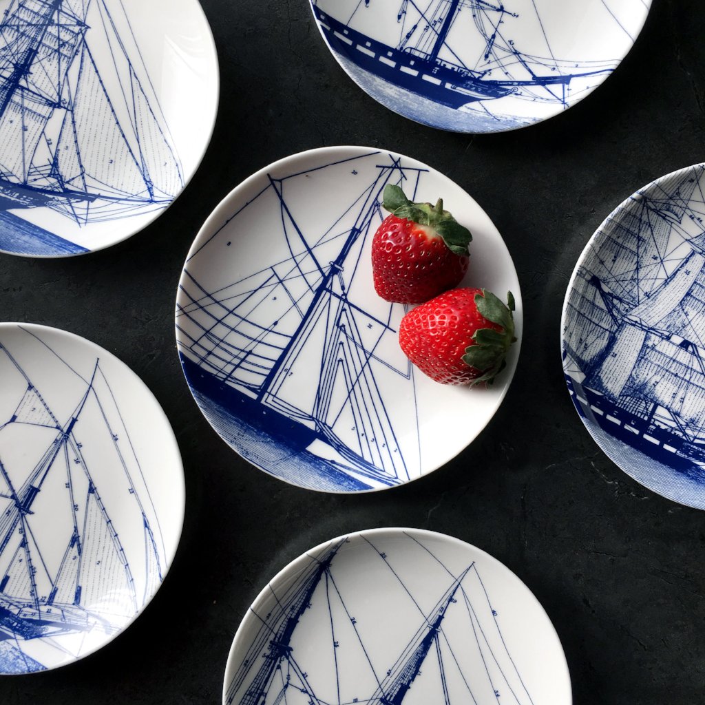 Small plates with vintage illustrations of blue ships arranged on a dark surface, featuring two fresh strawberries on one of the Caskata Artisanal Home Rigging Small Plates.
