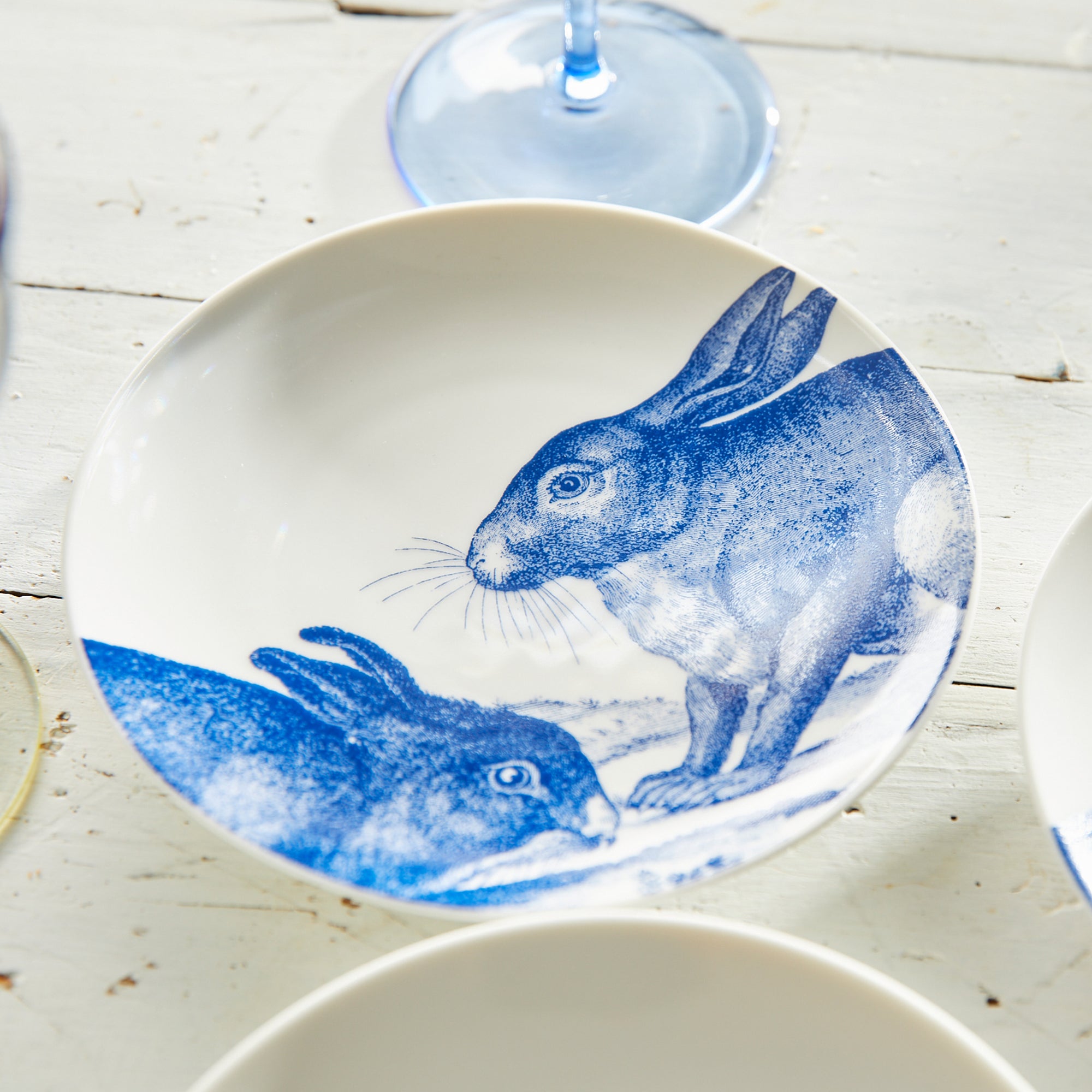 Four high-fired porcelain plates featuring blue illustrations of rabbits in various poses, called Bunnies Small Plates by Caskata Artisanal Home.