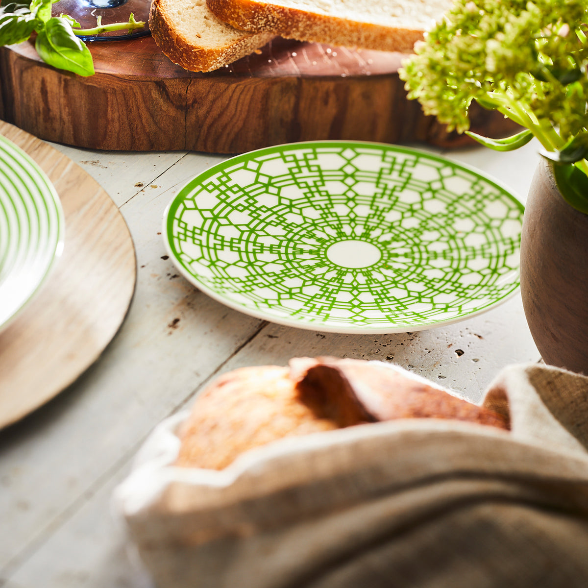 A green and white patterned Newport Verde Small Plate from the Caskata Artisanal Home collection sits on a wooden table next to a loaf of bread, sliced bread, and a pot of green flowers. This heirloom-quality dinnerware adds charm and elegance to any meal.