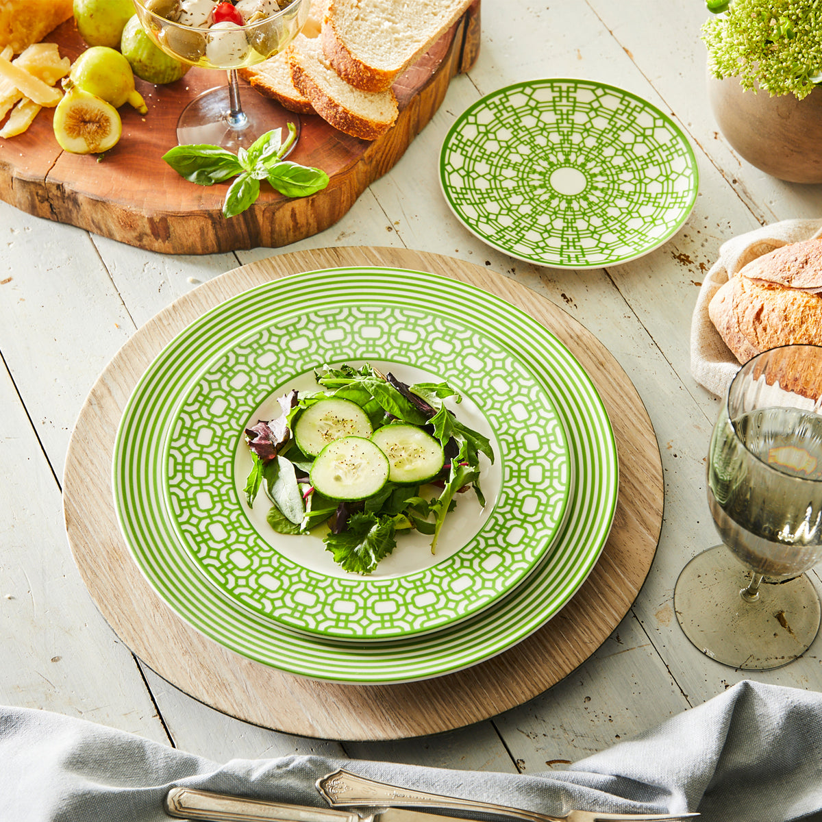 Newport Verde Small Plates from Caskata Artisanal Home, part of our Newport Garden Gate collection of premium porcelain, are stacked on a wooden charger holding a mixed green salad with cucumber slices. Nearby are bread, figs, a drink in a wine glass, and a small plant. This heirloom-quality dinnerware adds elegance to any meal.