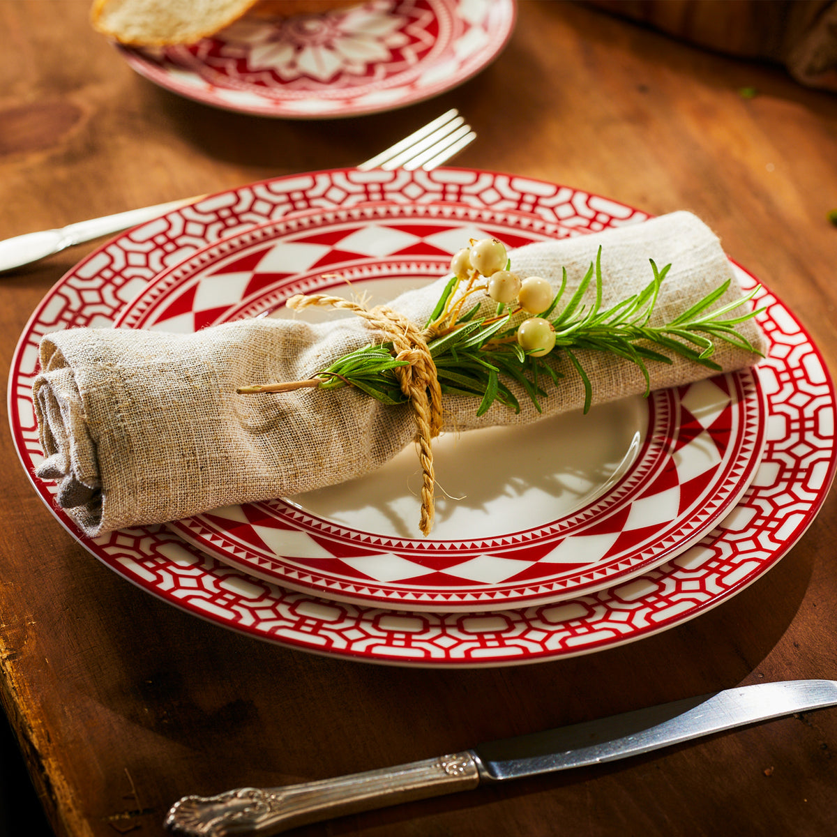 A red and white high-fired porcelain **Fez Crimson Rimmed Salad Plate** by **Caskata Artisanal Home** with a beige cloth napkin tied with twine and garnished with rosemary and white berries, set on a wooden table with a fork, knife, and slice of bread in the background.